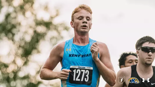 Kent State men's cross country runner in a light blue singlet with bib number 1273 runs with his mouth slightly open, while two competitors, one wearing sunglasses, follow close behind him.