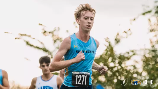 Kent State men's cross country runner in a light blue singlet with bib number 1271 runs with a determined look, his hair tousled, while a runner in a white and blue uniform trails behind him.