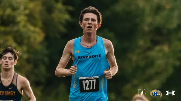 Kent State men's cross country runner in a light blue singlet with bib number 1277 runs toward the camera with his fists clenched and a silver chain around his neck, while a competitor in a navy and gold uniform trails behind him. Green trees fill the background.
