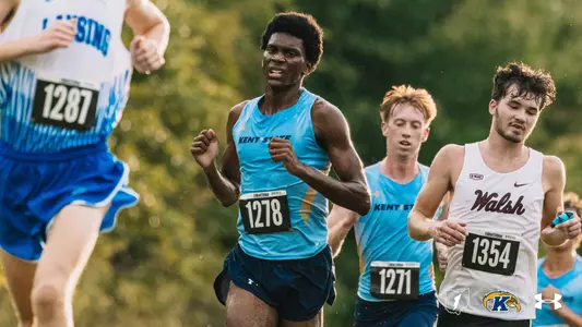Kent State men's cross country runner in a light blue singlet with bib number 1278 leads a pack on a grassy course, with a teammate wearing bib number 1271 close behind him. A runner in a white and blue uniform runs in the foreground, and a runner in a white Walsh uniform runs beside the teammate.