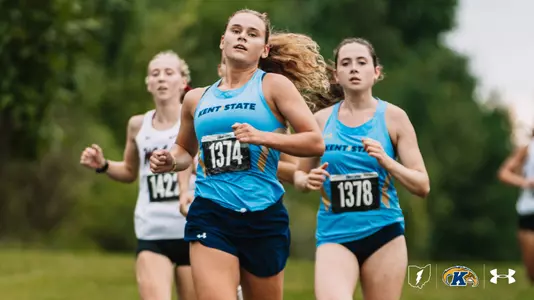 Two Kent State women's cross country runners in light blue singlets, bib numbers 1374 and 1378, run side by side down a grassy course, with a runner in a white uniform close behind them.
