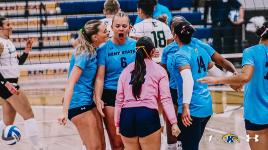 Kent State volleyball players in light blue jerseys celebrate in a huddle near the net, with No. 6 shouting in excitement while teammates cheer around her. A player in a pink long-sleeve shirt stands with her back to the camera, and a No. 14 teammate stands at right. A ball bounces in the lower left, and an opponent in white and green stands behind the net.
