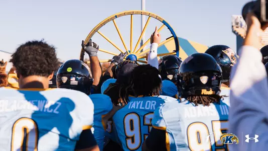 Kent State football players in light blue uniforms celebrating while holding up the Wagon Wheel trophy.