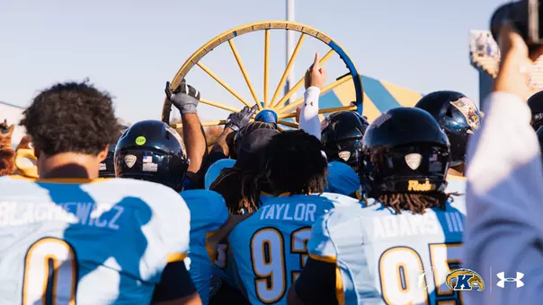 Kent State football players in light blue uniforms celebrating while holding up the Wagon Wheel trophy.