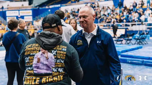 A man in a navy Kent State Gymnastics jacket smiling while talking to someone wearing a tribute shirt honoring head coach Brice Biggin.