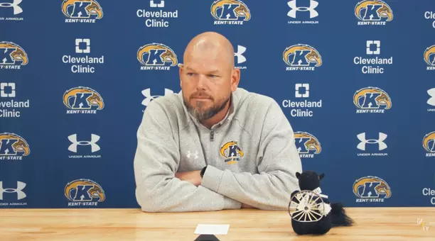 A bald man with a beard wearing a gray Kent State pullover seated at a desk during a press conference in front of a blue Kent State backdrop, next to a small black squirrel plush holding a miniature wagon wheel.