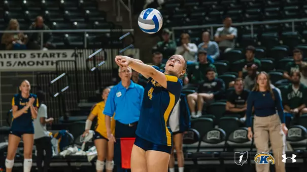 Kent State volleyball player #4, in a navy and gold uniform, looks up at the ball with her arms extended in a forearm pass during a match at Ohio, with teammates standing on the sideline behind her.