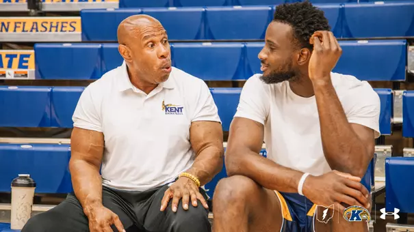 A Kent Basketball staff member in a white polo chats with a smiling man in a white t-shirt on the arena bleachers.
