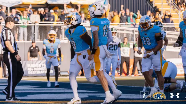 American football players wearing light blue jerseys and white pants celebrate a play on the field during a game as a referee looks on from the left.