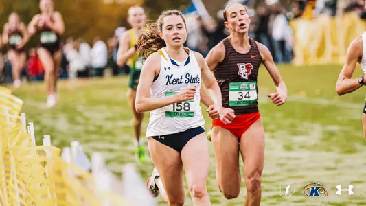 Kent State’s Alexis Tracy, wearing bib 158 and a white Kent State jersey with navy shorts, runs in a cross country race with a focused expression while Bowling Green’s Boone, bib 34, runs close behind her in a brown striped top and orange shorts. Other runners are blurred in the background, with yellow course fencing along the left and spectators lining the grass course.