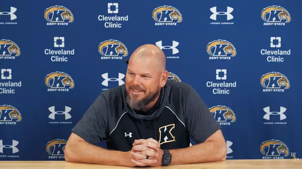 A bald man with a beard wearing a gray Kent State shirt sits with clasped hands during a press conference in front of a blue Kent State backdrop.