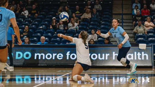 A volleyball player in a white jersey with the number 5 lunges forward on one knee with both arms extended low to dig the ball, which is just in front of her. Behind her, a Kent State teammate in a light blue jersey with the number 14 closes in, watching the ball. Another light blue-clad player is partially visible at the left edge of the frame. Fans sit in navy arena seats in the background, and an LED board along the court reads “DO more for your health.
