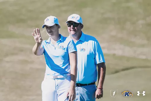 Veronika Kedronová (left) looks toward the green and raises her hand while head coach Casey VanDamme (right) stands beside her, coaching her through a putt; both wear light blue Kent State polos and white caps on the course.