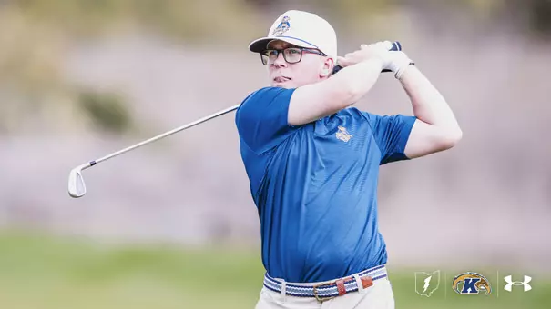 Liam Curtis follows through on an iron shot during a Kent State men's golf practice round. Curtis wears a blue Kent State polo with the Fearless Flash logo, a white cap, glasses, and a white golf glove. Logos for Kent State athletics, the Flashes, and Under Armour appear in the bottom right corner.