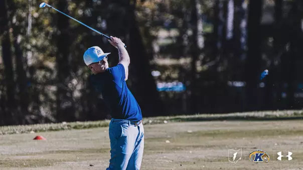 Kent State first-year Liam Curtis follows through on an iron shot during a practice round, wearing a blue polo and white Kent State cap with trees in the background.
