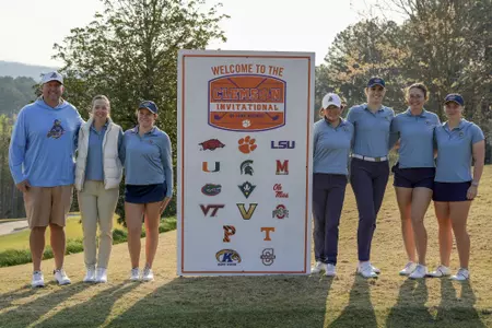 Kent State women's golf team poses on the course next to the Clemson Invitational on Lake Keowee welcome sign, which displays the logos of all participating teams. From left to right: head coach Casey VanDamme, assistant coach Manuela Carbajo Ré, Isabella Goyette, Leon Takagi, Veronika Kedronová, Petra Babicová and Gracie Larsen. The team is wearing Kent State light blue with trees and a hazy sky in the background.