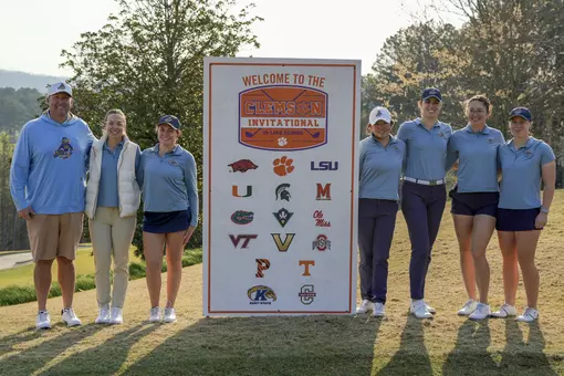 Kent State women's golf team poses on the course next to the Clemson Invitational on Lake Keowee welcome sign, which displays the logos of all participating teams. From left to right: head coach Casey VanDamme, assistant coach Manuela Carbajo Ré, Isabella Goyette, Leon Takagi, Veronika Kedronová, Petra Babicová and Gracie Larsen. The team is wearing Kent State light blue with trees and a hazy sky in the background.