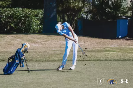 Kent State’s Veronika Kedronová swings through an iron shot from the fairway during a sunny round of play, taking a divot as dirt lifts into the air. Her Kent State golf bag stands beside her on the left, with trees and greenery in the background.