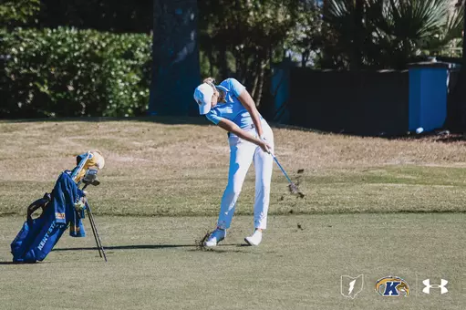 Kent State’s Veronika Kedronová swings through an iron shot from the fairway during a sunny round of play, taking a divot as dirt lifts into the air. Her Kent State golf bag stands beside her on the left, with trees and greenery in the background.
