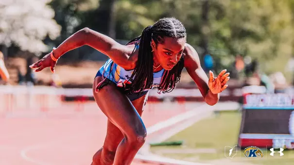 Kent State track and field athlete Miranna Atanmo bursts out of the starting blocks, leaning forward in full acceleration with arms driving and braided hair flowing behind her. Hurdles are visible in the background.