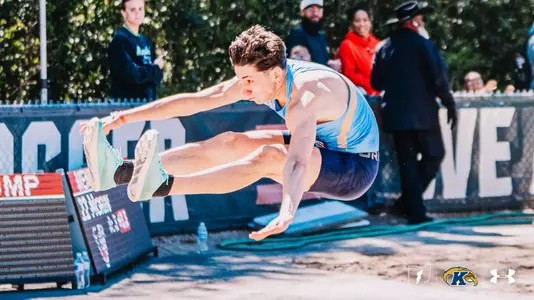 Kent State track and field athlete Luigi Case soars through the air in the long jump, extending his legs forward and reaching toward his feet in a hitch-kick position above the sand pit.