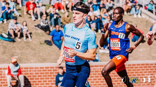 Kent State track and field athlete Dylan McBride, wearing an Under Armour headband and bib number 5, competes at the Raleigh Relays, running alongside a Clemson athlete in front of a crowded hillside and stadium crowd.