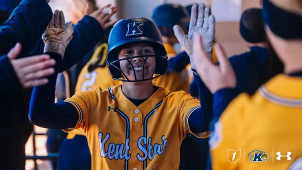 Kent State University softball player #1 Brooke Aberle, wearing a gold 'Kent State' jersey and navy blue batting helmet with face guard, celebrates in the dugout with a wide smile and both arms raised to exchange high-fives with surrounding teammates. Teammates in gold and navy uniforms are visible in the background. The Kent State logo, Ohio outline, and Under Armour logo appear in the lower right corner
