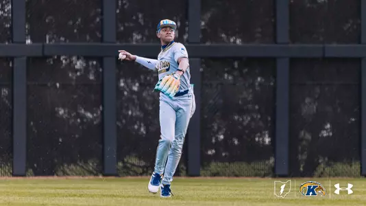 A Kent State University baseball player in a gray road uniform with ‘Flashes’ across the chest winds up to throw the ball from the outfield. The player wears a blue cap, a colorful glove, and blue cleats. A green warning track and dark outfield wall are visible in the background. The Kent State ‘K’ logo and Under Armour logo appear in the bottom right corner.