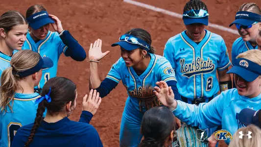 Kent State softball player Aaralyn Nogay (#11) grins broadly as she runs through a celebratory tunnel of high-fives from her teammates during pre-game introductions. Nogay, wearing a navy blue visor with reflective sunglasses pushed up on the brim, reaches both hands out to slap hands with teammates gathered around her. The team is dressed in light blue Kent State softball uniforms with navy and gold accents and the cursive 'Kent State' wordmark across the chest. Several players wear navy 'Kent State' visors, and teammate #2 is visible in the background. The scene takes place on a red clay infield, capturing an energetic and joyful team moment before the game.