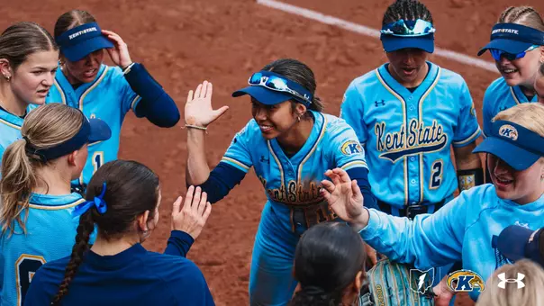 Kent State softball player Aaralyn Nogay (#11) grins broadly as she runs through a celebratory tunnel of high-fives from her teammates during pre-game introductions. Nogay, wearing a navy blue visor with reflective sunglasses pushed up on the brim, reaches both hands out to slap hands with teammates gathered around her. The team is dressed in light blue Kent State softball uniforms with navy and gold accents and the cursive 'Kent State' wordmark across the chest. Several players wear navy 'Kent State' visors, and teammate #2 is visible in the background. The scene takes place on a red clay infield, capturing an energetic and joyful team moment before the game.