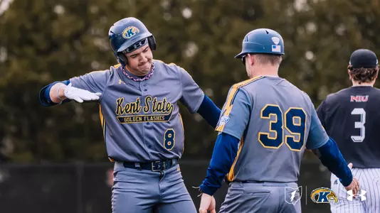 Kent State Golden Flashes baseball player wearing No. 8 exchanges a fist bump with a Kent State coach wearing No. 39 near the base paths. Both are in gray road uniforms with ‘Kent State Golden Flashes’ lettering in blue and gold. A Northern Illinois player wearing No. 3 is visible in the background. The Kent State ‘K’ logo and Under Armour logo appear in the bottom right corner.