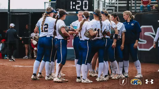 A group of Kent State University softball players in white jerseys and blue pants gather in a huddle on the infield dirt during a game. Several players wear blue headbands and have braided hair. A coach in a navy blue Kent State jacket stands to the right. A digital scoreboard displaying "1:20" is visible in the background. The Kent State "K" logo and Under Armour logo appear in the bottom right corner.
