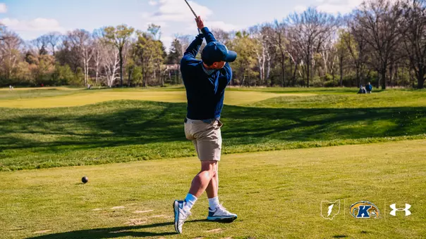 A Kent State men's golfer completes a full follow-through on a tee shot, his arms extended high above his head and his back turned to the camera as he watches the ball travel down the fairway. The golfer wears a navy blue quarter-zip pullover, khaki shorts, white socks, and white and blue golf shoes with a navy cap. The ball is visible in the lower left of the frame, just off the tee box. A wide, sun-lit fairway stretches ahead with trees lining both sides and a partly cloudy sky above. Several other golfers are visible in the distant background. The Kent State, Ohio Lightning Bolt, and Under Armour logos appear in the bottom right corner.