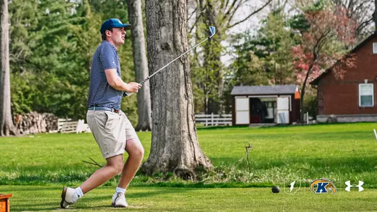 Kent State men's golfer Christoffer Leuenhagen follows through on an iron shot, his body turned and weight shifted forward in his follow-through as he watches the ball in flight. Leuenhagen wears a blue and white striped polo, khaki shorts, white golf shoes, and a navy blue cap. The shot takes place on a lush green fairway with large trees, a white fence, and a red brick building visible in the background. The Kent State, Fearless Flash, and Under Armour logos appear in the bottom right corner.