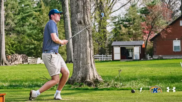 Kent State men's golfer Christoffer Leuenhagen follows through on an iron shot, his body turned and weight shifted forward in his follow-through as he watches the ball in flight. Leuenhagen wears a blue and white striped polo, khaki shorts, white golf shoes, and a navy blue cap. The shot takes place on a lush green fairway with large trees, a white fence, and a red brick building visible in the background. The Kent State, Fearless Flash, and Under Armour logos appear in the bottom right corner.