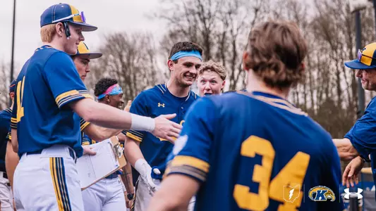 A Kent State baseball player wearing a light blue headband smiles broadly as he is greeted by teammates in the dugout after scoring a run, exchanging handshakes and fist bumps with players in navy and gold Under Armour uniforms. Jersey number 34 is visible in the foreground as several teammates and a coach look on with excitement, with bare winter trees visible in the background on an overcast day.