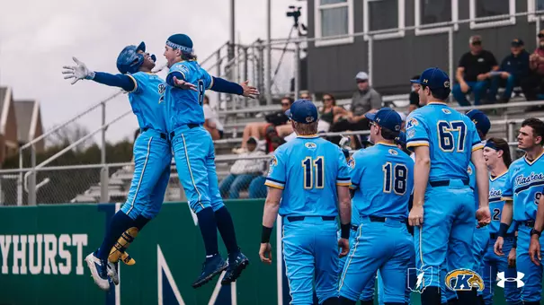 Two Kent State Golden Flashes baseball players leap into the air for a jubilant chest bump celebration near the dugout after a big play, both wearing the team's powder blue "Flashes" alternate uniforms. Several teammates in jerseys numbered 11, 18, and 67 look on from the dugout steps, while fans in the bleachers watch in the background. The green outfield wall with partial "yhurst" lettering is visible, indicating the game is being played at an opponent's home field.