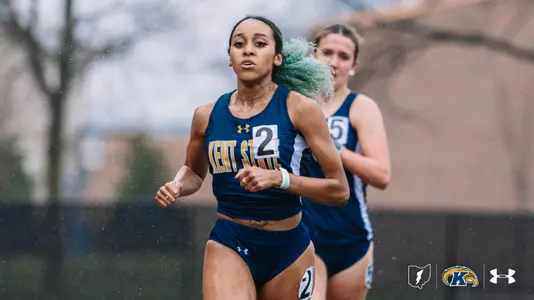 Kent State University track and field athlete Camille Dunifer, wearing bib number 2, races in a distance event during an outdoor meet, leading a competitor in a navy blue uniform. The Golden Flashes runner wears the navy blue and gold Kent State uniform with an Under Armour logo. Rain is visible in the background. The Kent State Athletics logo and Under Armour wordmark appear in the lower right corner.