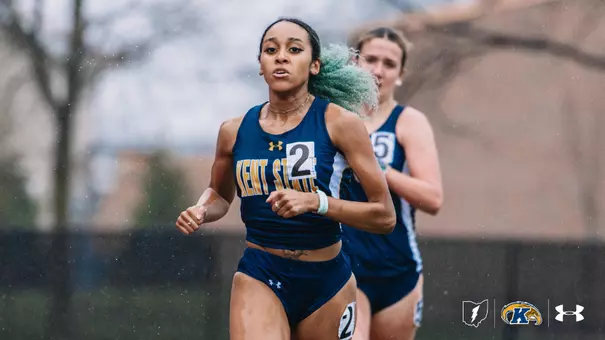 Kent State University track and field athlete Camille Dunifer, wearing bib number 2, races in a distance event during an outdoor meet, leading a competitor in a navy blue uniform. The Golden Flashes runner wears the navy blue and gold Kent State uniform with an Under Armour logo. Rain is visible in the background. The Kent State Athletics logo and Under Armour wordmark appear in the lower right corner.