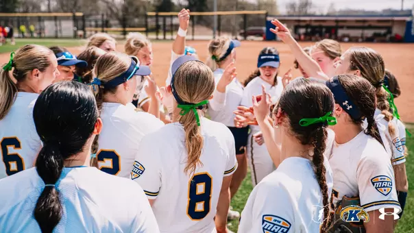 The Kent State softball team huddles together on the field, hands raised and celebrating, wearing white home uniforms with navy and gold MAC Conference patches. Players sport green ribbon hair accessories, with the softball diamond visible in the background.
