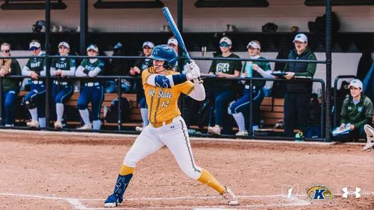 "Kent State University softball player Ashlyn Porter (#12) stands in a left-handed batting stance at home plate, holding a blue bat. She is dressed in the team's gold and white Under Armour uniform with a blue helmet and leg guard. In the background, players and staff from the opposing team — dressed in green — watch from the dugout. The Kent State and Under Armour logos are visible in the lower right corner."