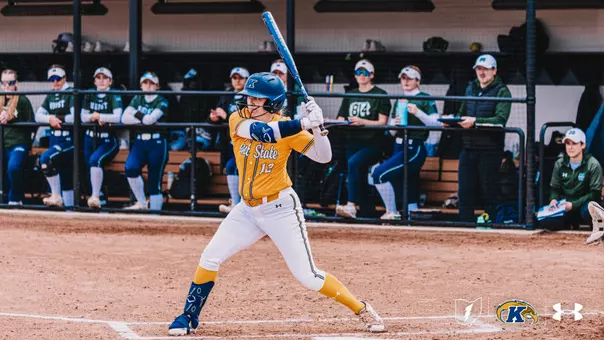 "Kent State University softball player Ashlyn Porter (#12) stands in a left-handed batting stance at home plate, holding a blue bat. She is dressed in the team's gold and white Under Armour uniform with a blue helmet and leg guard. In the background, players and staff from the opposing team — dressed in green — watch from the dugout. The Kent State and Under Armour logos are visible in the lower right corner."