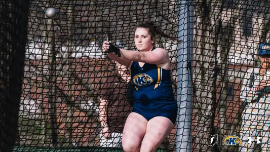 Kent State University track and field athlete Kaylee Carlisle winds up during a hammer throw attempt, gripping the handle with both hands as the implement blurs overhead. She wears a navy blue and gold Kent State uniform featuring the Golden Flashes logo. A safety cage surrounds the throwing circle. The Kent State Athletics logo and Under Armour wordmark appear in the lower right corner.