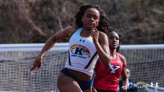 Kent State University sprinter Nia Massey drives hard out of her lane during an outdoor track meet, wearing a white Kent State uniform featuring the Golden Flashes logo. A competitor in a red Youngstown State uniform races alongside her. The Kent State Athletics logo and Under Armour wordmark appear in the lower right corner.