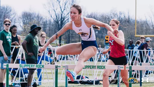 Kent State University track and field athlete Keeli Dunaway clears a hurdle at the Slippery Rock outdoor track meet, wearing a white Kent State uniform with the Golden Flashes logo. A Youngstown State competitor in a red uniform races alongside her. Spectators line the infield in the background. The Kent State Athletics logo and Under Armour wordmark appear in the lower right corner.