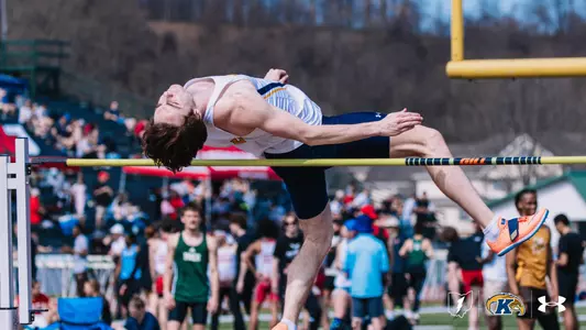 Kent State University track and field athlete Cooper DeFrange arches over the bar using the Fosbury Flop technique during an outdoor meet at Slippery Rock. He wears a white and navy Kent State uniform. A large crowd of spectators and a football goal post are visible in the background. The Kent State Athletics logo and Under Armour wordmark appear in the lower right corner.