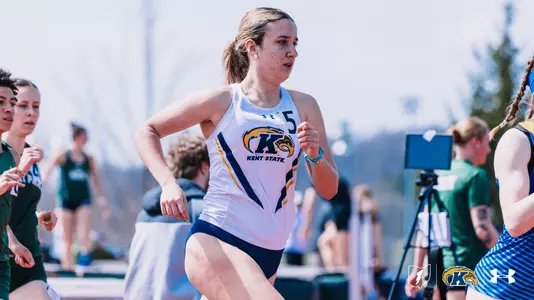 Kent State University distance runner Ana Fernandez Moiron, wearing bib number 5, competes in an outdoor track event at Slippery Rock, dressed in a white Kent State uniform featuring the Golden Flashes logo. Competitors from other programs and meet officials are visible in the background. The Kent State Athletics logo and Under Armour wordmark appear in the lower right corner.