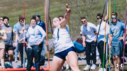 Kent State University track and field athlete Kristen Jay releases a javelin throw during an outdoor meet at Slippery Rock, wearing a white and navy Kent State uniform with the Golden Flashes logo. Javelins planted in the ground and a crowd of spectators are visible in the background. The Kent State Athletics logo and Under Armour wordmark appear in the lower right corner.