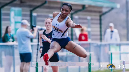 Kent State University track and field athlete Destiny Nash clears a hurdle during an outdoor track meet at Slippery Rock, wearing a white Kent State uniform featuring the Golden Flashes logo. A competitor in a dark uniform trails behind her. Officials and spectators are visible in the background. The Kent State Athletics logo and Under Armour wordmark appear in the lower right corner.