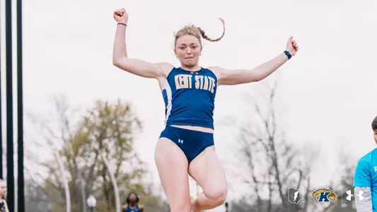 Kent State University track and field athlete Ally Schindler soars through the air during a long jump attempt at an outdoor meet, arms raised and knees lifted. She wears the navy blue and gold Kent State uniform. The Kent State Athletics logo and Under Armour wordmark appear in the lower right corner.